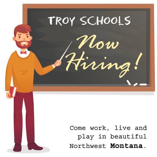 An image of a smiling bearded man pointing to a chalkboard that reads "Troy Schools Now Hiring" with the caption "Come work, live and play in beautiful Northwest Montana."
